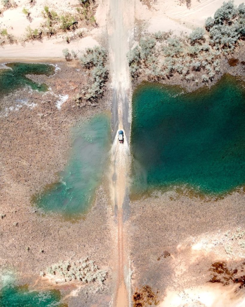 A 4WD splashes through water on Gibb River Road, surrounded by red earth and outback vegetation—classic rugged terrain for road trips in Australia.