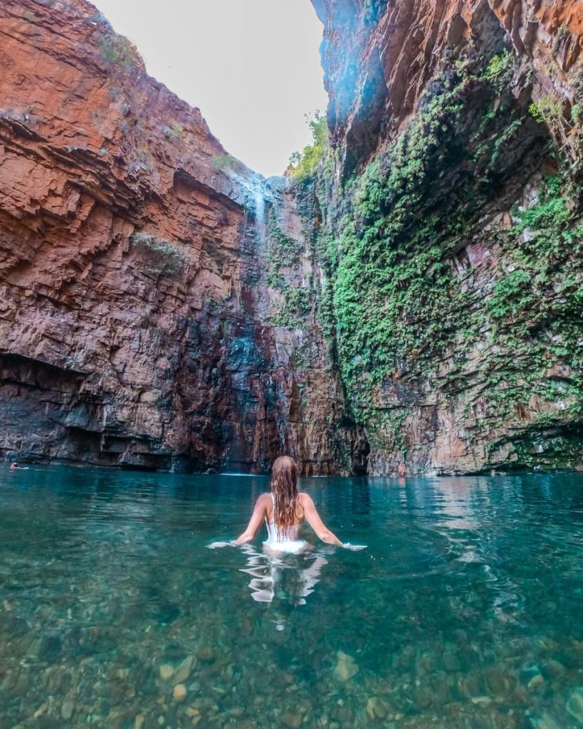 A woman swims beneath a thin waterfall cascading down the red cliffs of Emma Gorge in Western Australia—pure magic on Kimberley road trips in Australia.
