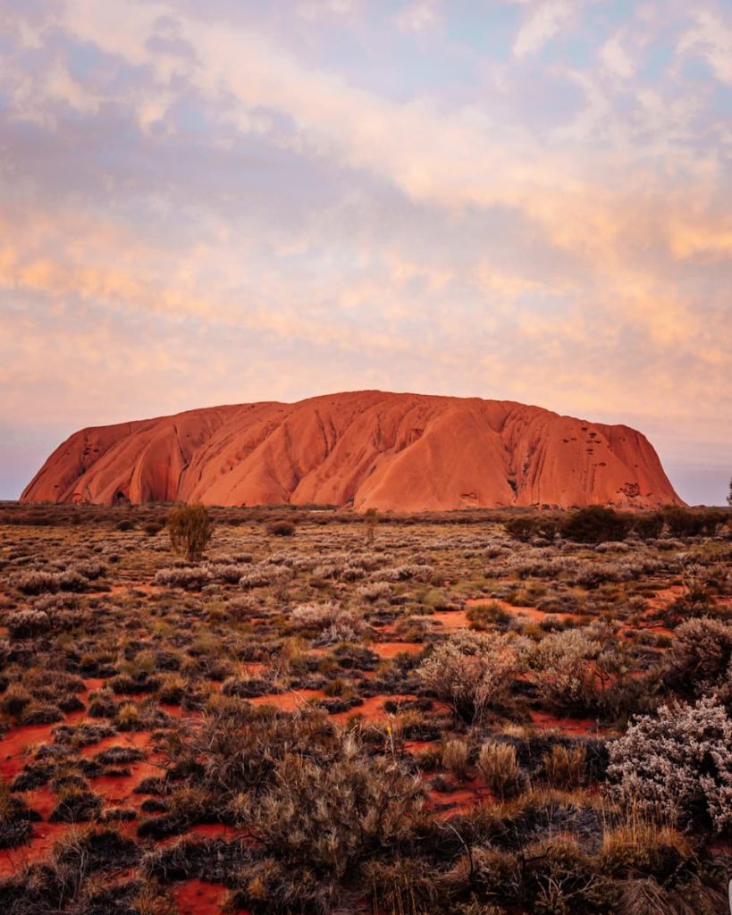 Uluru at sunrise with soft pastel skies above and red desert landscape in the foreground, one of the most iconic stops on road trips in Australia’s Northern Territory.