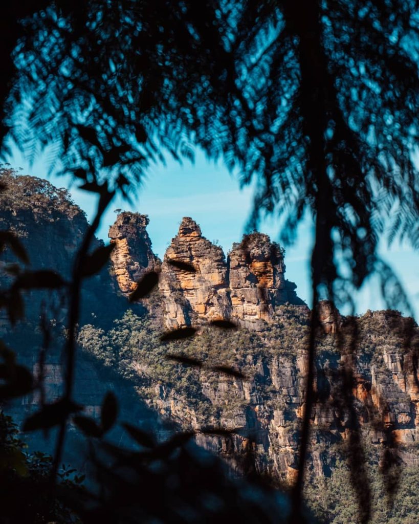 The Three Sisters rock formation framed by fern leaves against a clear sky, a must-see landmark on a Blue Mountains weekend itinerary.