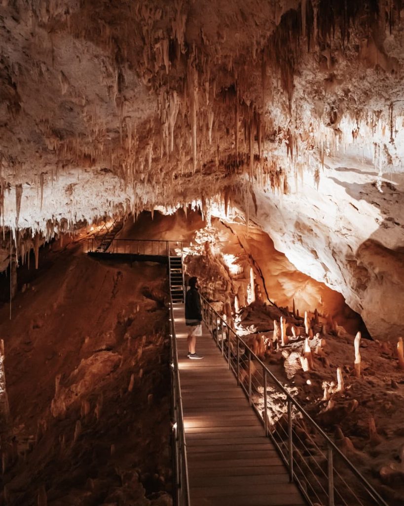 A person stands on a wooden walkway inside a limestone cave in Margaret River, surrounded by stalactites and warm lighting, a hidden gem on road trips in Australia.