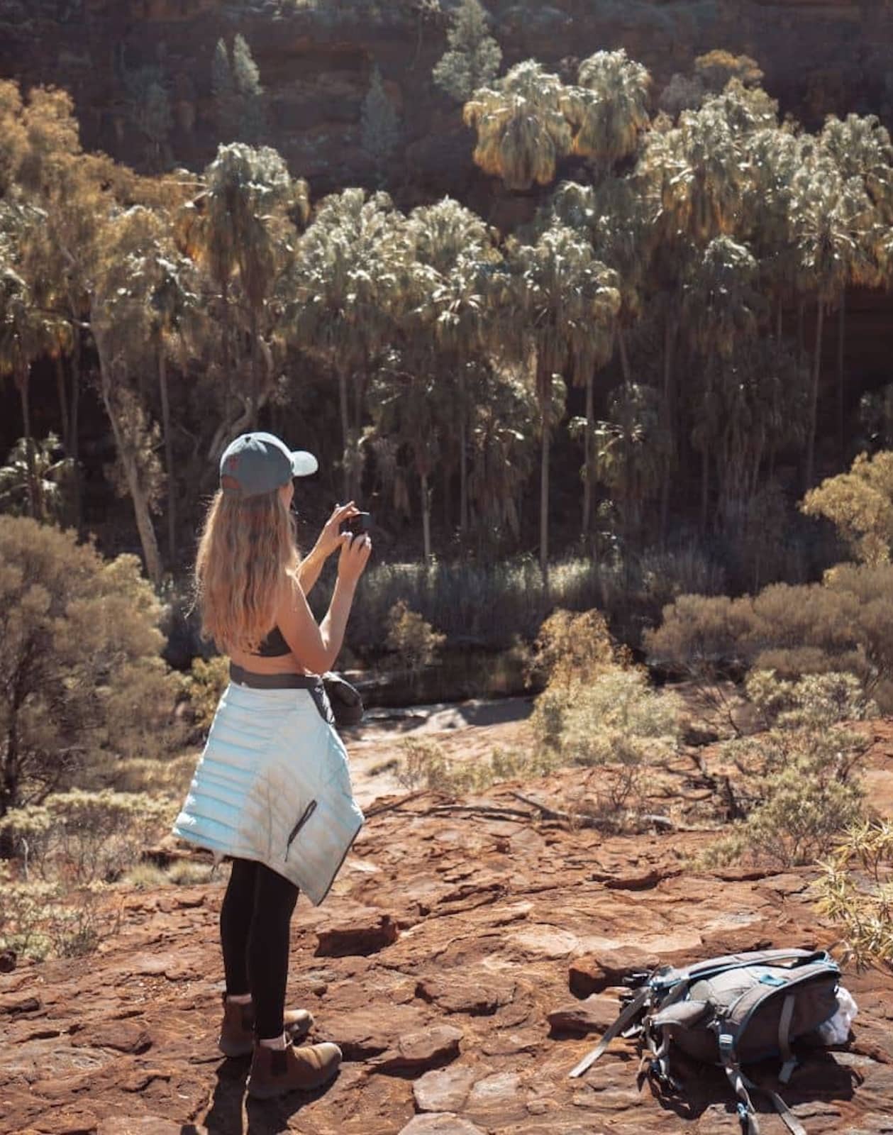 A woman in hiking gear stands on a rocky ledge photographing a lush oasis of tall palm trees nestled in a gorge in the West MacDonnell Ranges. Her backpack lies on the ground nearby under the warm sunlight.