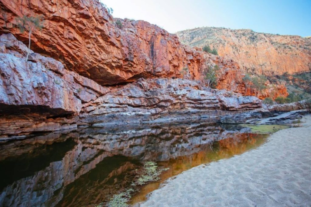 The red rock walls of Ormiston Gorge in the Northern Territory, Australia, reflected in a calm pool of water surrounded by sandy banks.