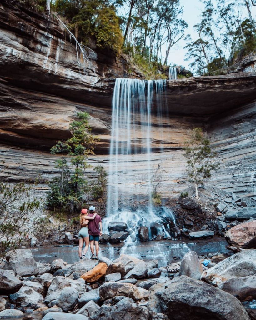 A couple stands arm in arm before Victoria Falls, watching water plunge from a rocky ledge—an intimate and rewarding pause on any Blue Mountains weekend itinerary.