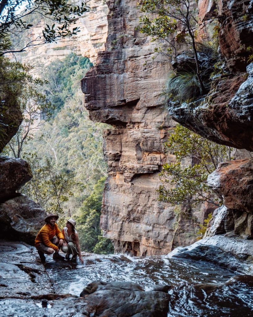 A couple kneels at the edge of a rocky stream near a cliff drop at the Leura Cascades infinity pool, tucked away along a hidden Blue Mountains weekend route.