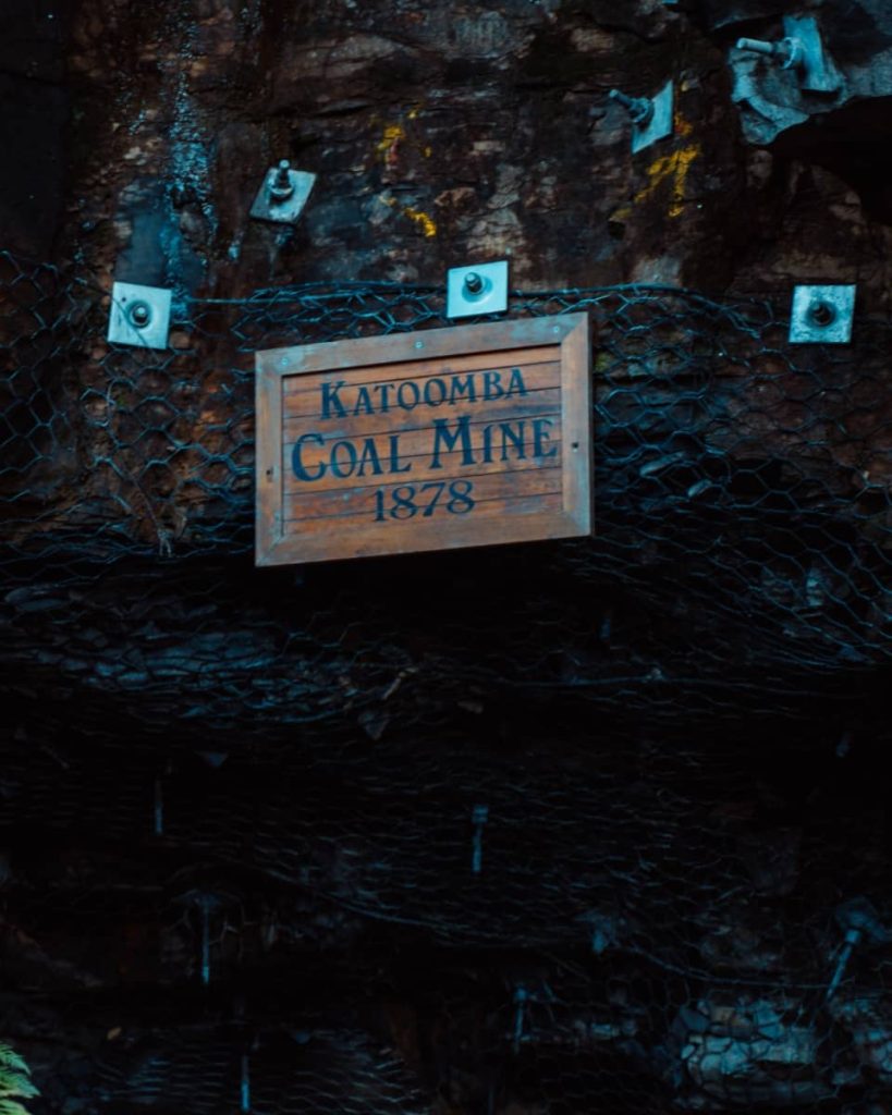 A wooden sign reading “Katoomba Coal Mine 1878” is mounted against a rock wall, showcasing a historic stop along Scenic World’s walkways and a cultural feature of the Blue Mountains weekend itinerary.