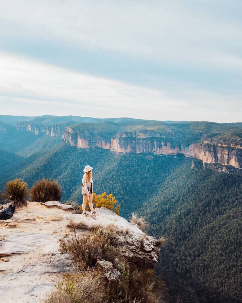 A woman in a white hat stands on the edge of a rocky cliff at Hanging Rock, overlooking dramatic cliffs and deep green valleys—one of the most striking spots on a Blue Mountains weekend itinerary.