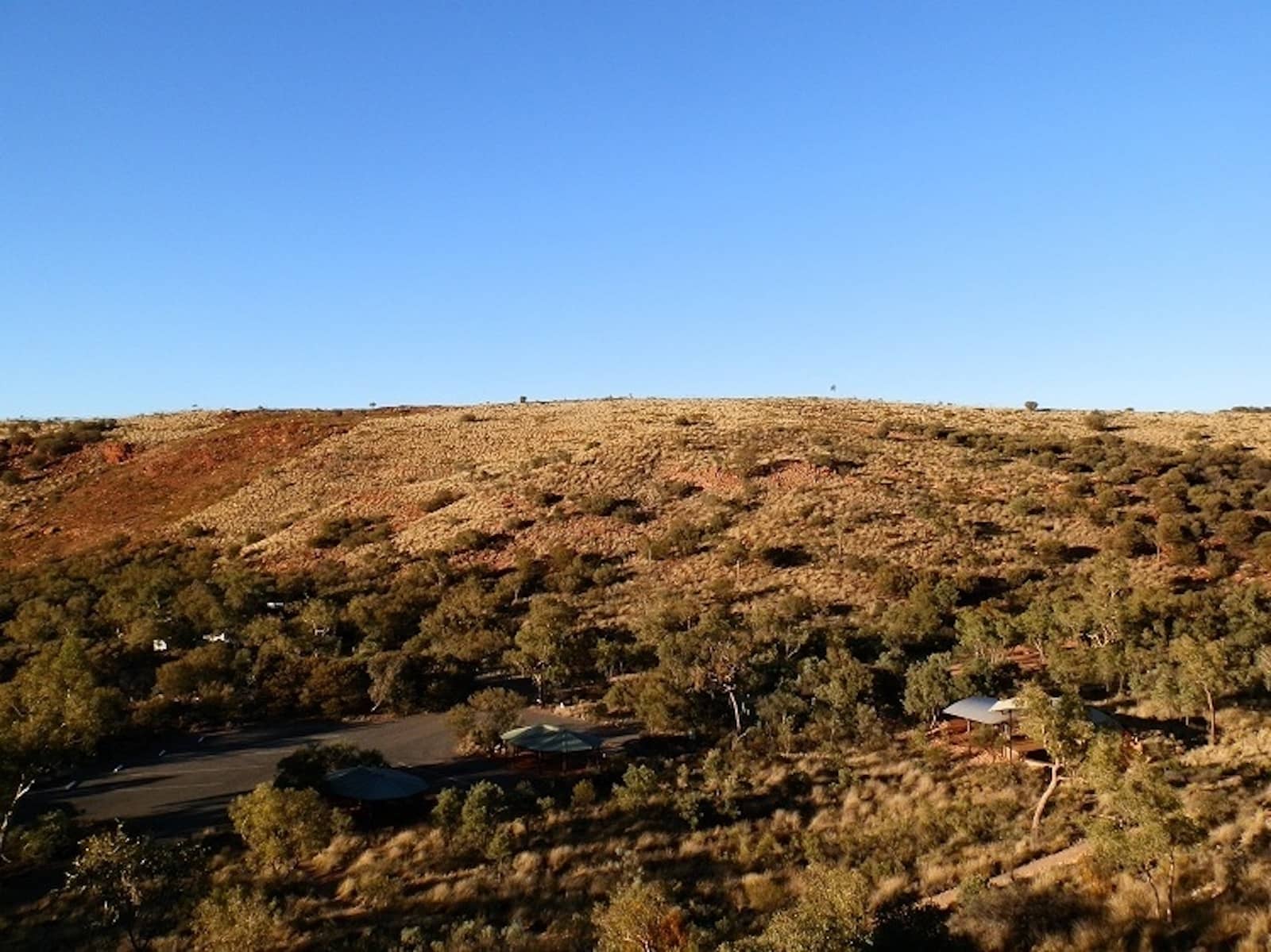 A view from a lookout trail shows a dry, grassy hillside dotted with sparse trees and a small campground with shaded shelters nestled at the base.