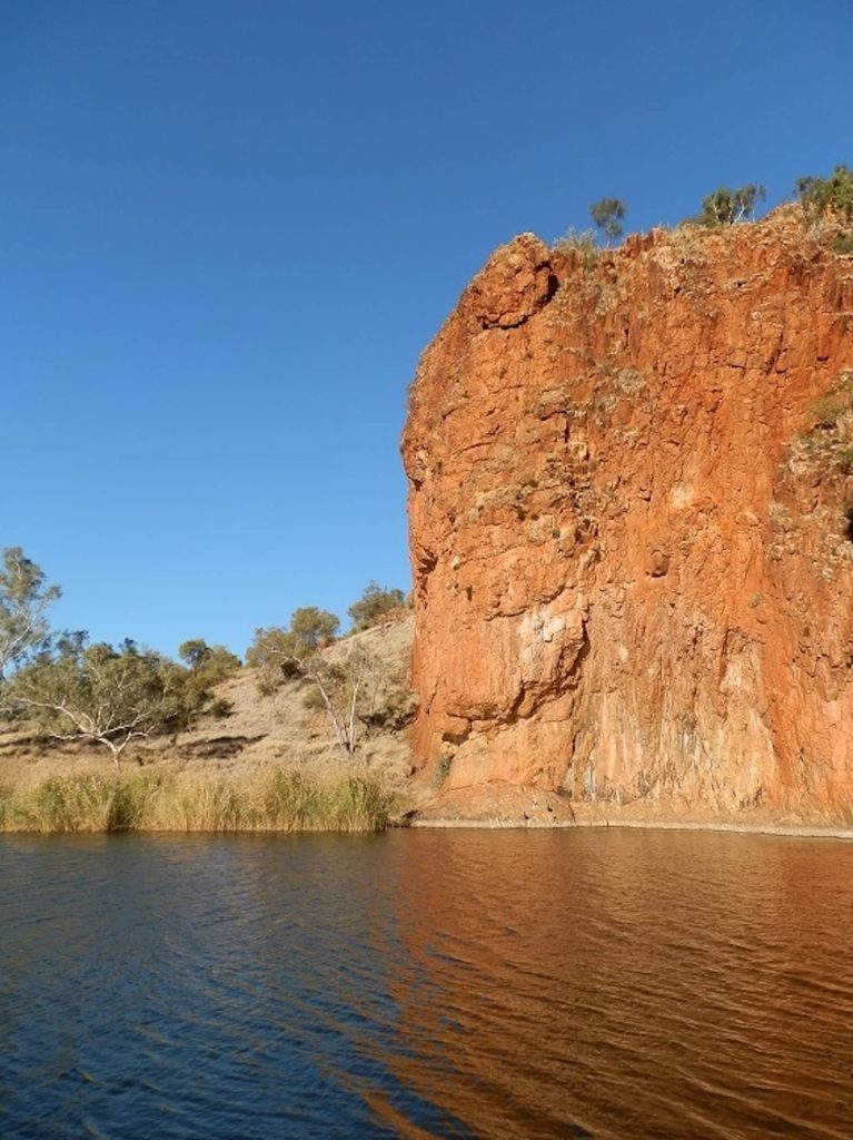 A towering red rock cliff rises dramatically from the edge of a calm waterhole, surrounded by dry grass, sparse trees, and a clear blue sky in the Glen Helen Gorge area.