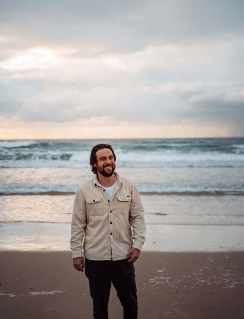 A man stands on the beach at the Gold Coast, smiling as the waves roll in behind him. The sky is overcast with hints of sunlight breaking through, casting a soft glow on the ocean. He wears a light-colored jacket and dark pants, enjoying the peaceful shoreline.