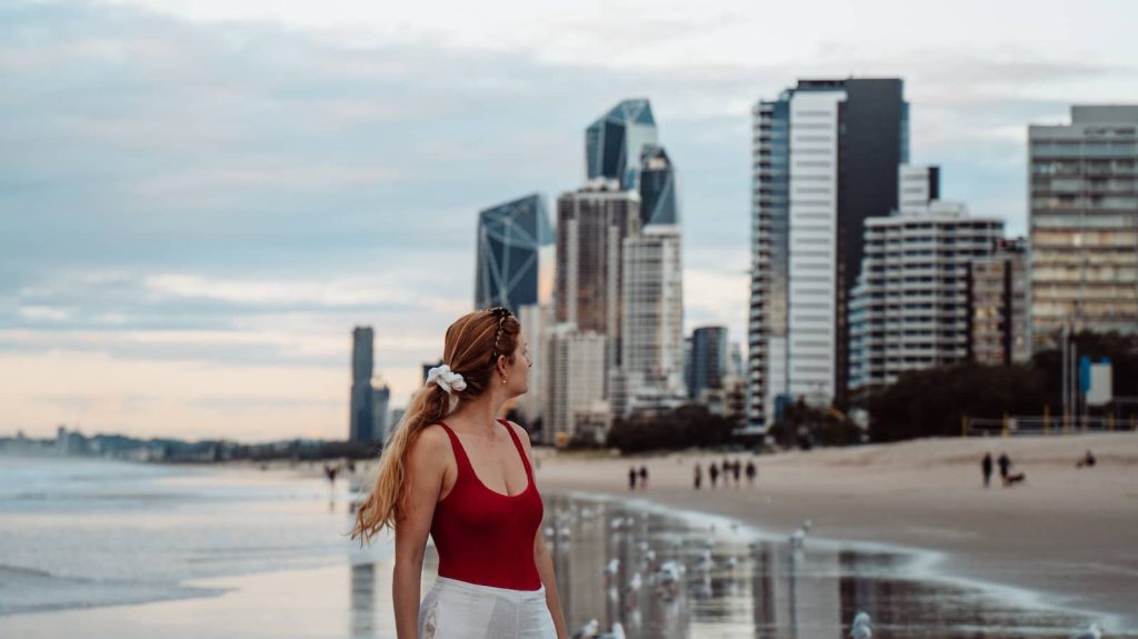 A woman in a red swimsuit and white shorts stands on Gold Coast beach at sunset, gazing at the modern skyline with glass skyscrapers in the background and seagulls along the shoreline.