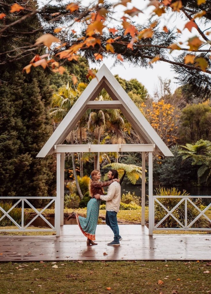 A couple stands under a charming white gazebo, surrounded by lush greenery and autumn foliage. The woman, dressed in a flowing blue skirt and brown leather jacket, playfully lifts one foot while holding onto her partner, who smiles back at her. The scene is framed by branches with golden leaves, adding a romantic touch.