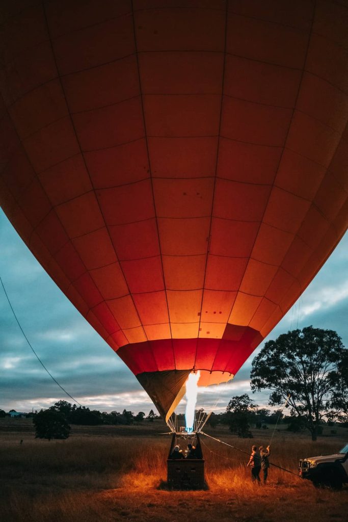 A hot air balloon is being inflated at dawn, with flames from the burner lighting up the inside of the balloon. Crew members hold onto the ropes while the balloon prepares for liftoff. The sky is a deep blue with hints of sunrise, and the field below is bathed in soft morning light.