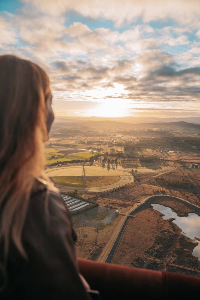 A woman gazes out over the vast countryside from a hot air balloon, bathed in the golden glow of sunrise. Below, a patchwork of farmland, winding waterways, and rolling hills stretch to the horizon, creating a breathtaking aerial view. The warm light highlights her long, wavy hair as she takes in the peaceful scenery.
