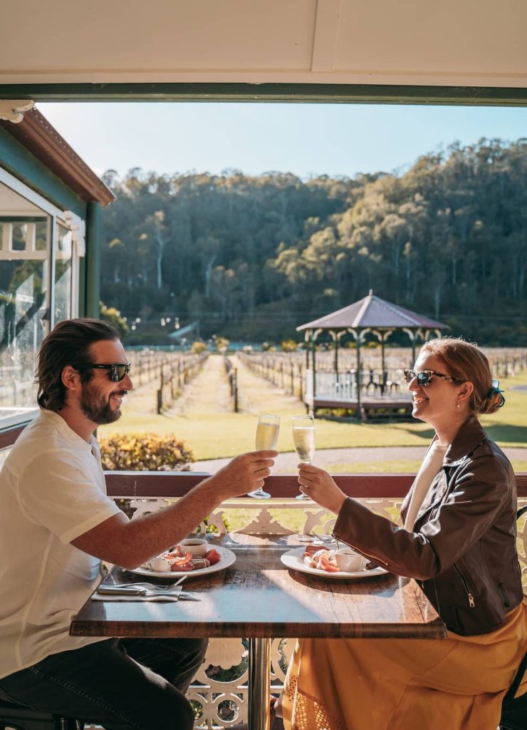 A couple enjoys breakfast with sparkling wine at an outdoor vineyard setting, with rows of grapevines and a gazebo in the background. The morning sunlight highlights their relaxed atmosphere as they toast with champagne glasses over a spread of fresh fruits and pastries.