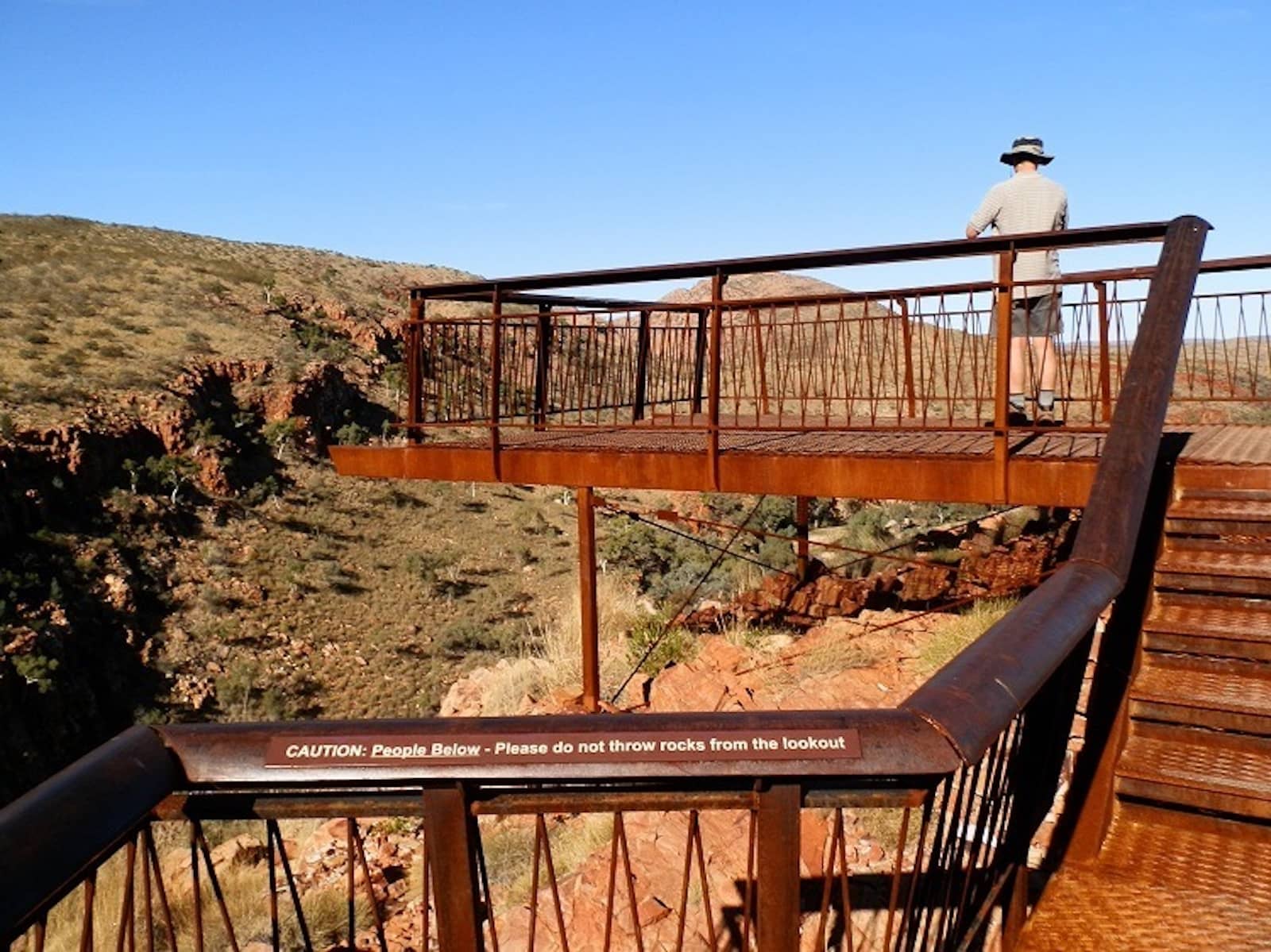 Elevated rust-colored metal lookout platform at Ghost Gum Lookout, with a person standing at the edge overlooking a rugged, grassy gorge. A warning sign reads, “CAUTION: People Below - Please do not throw rocks from the lookout.