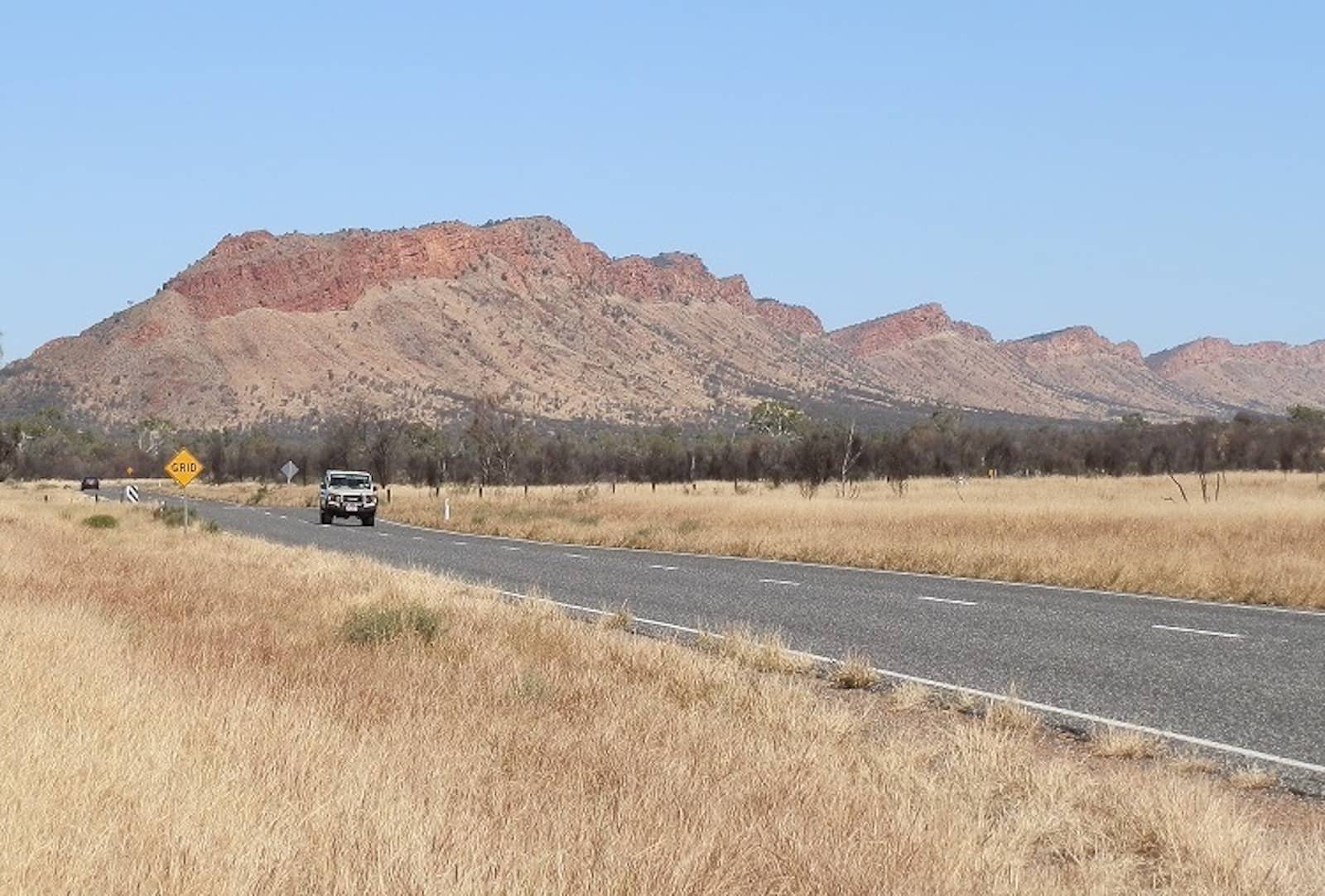 A 4WD vehicle drives along a paved road cutting through golden dry grasslands, with the rugged red ridges of the West MacDonnell Ranges rising in the background under a clear blue sky.