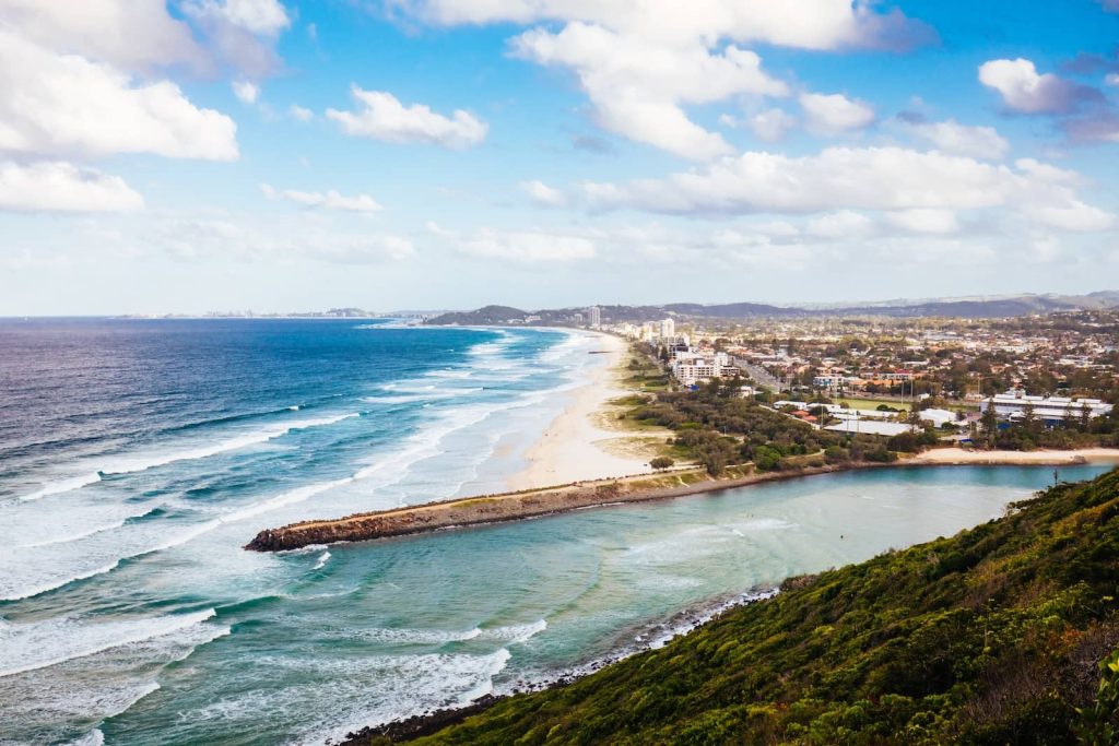 A stunning aerial view of Burleigh Head National Park, showcasing the coastline stretching towards the Gold Coast skyline. The turquoise ocean meets golden sandy shores, with waves rolling onto the beach. The greenery of the park contrasts with the urban landscape in the distance.