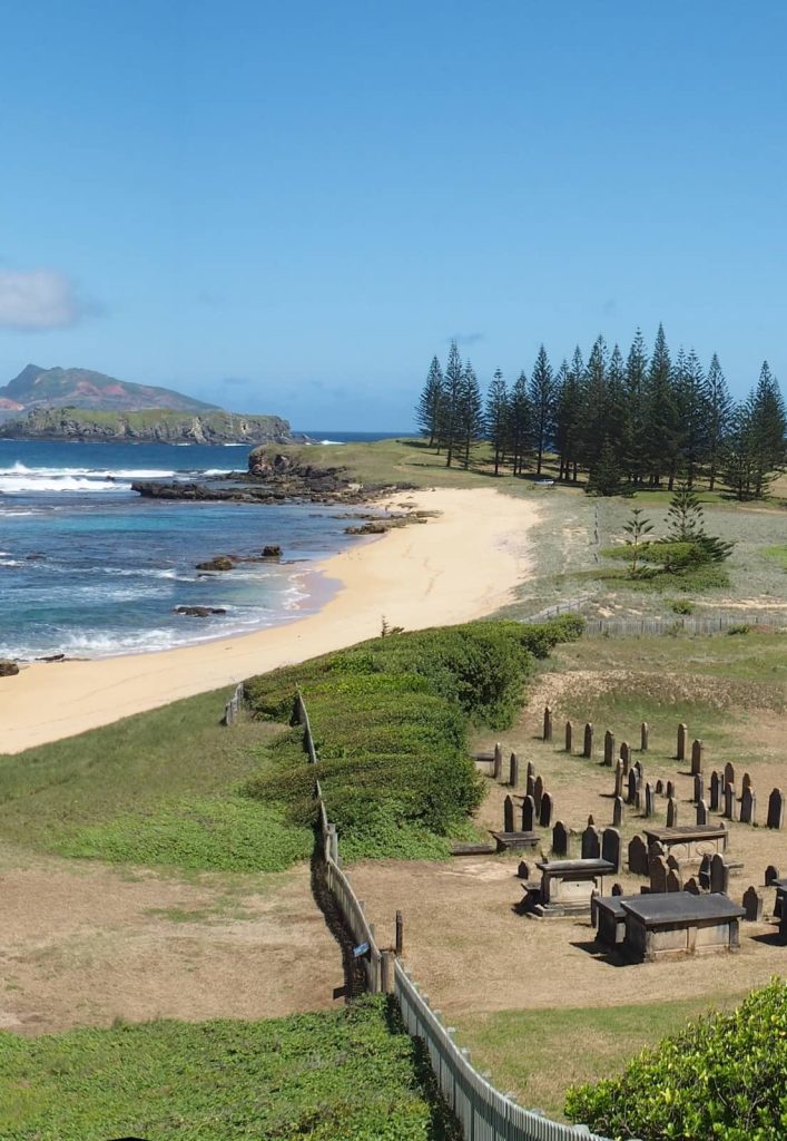 Scenic view of Cemetery Bay on Norfolk Island, featuring a historic graveyard near the shoreline, golden sand beach, rocky shoreline, and pine trees, with a distant island across the turquoise ocean.