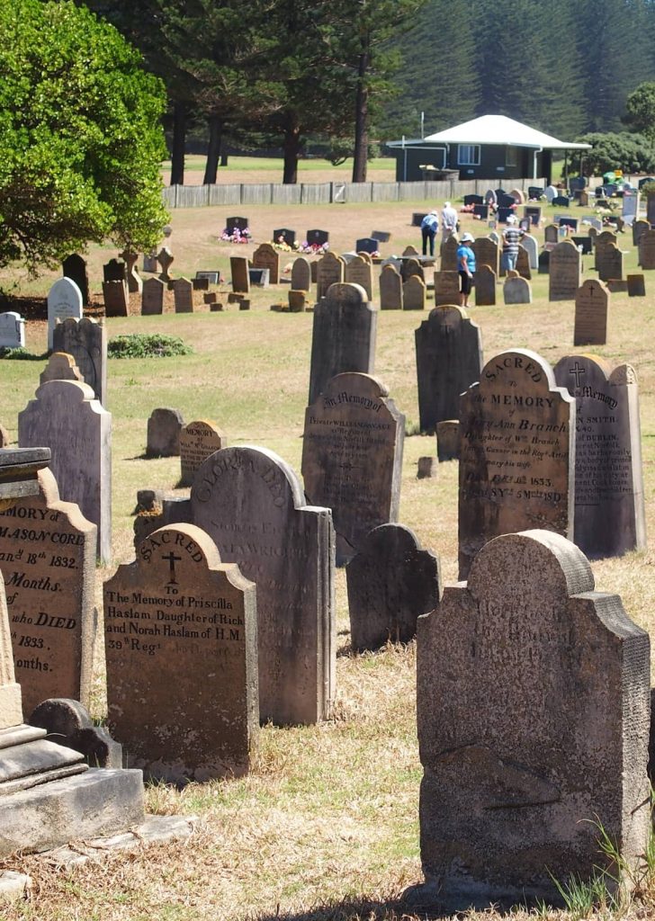 Close-up view of historic headstones in the Norfolk Island cemetery, with worn inscriptions and moss-covered markers under bright sunlight, while visitors explore the grounds in the distance near tall Norfolk pines and a green-roofed building.