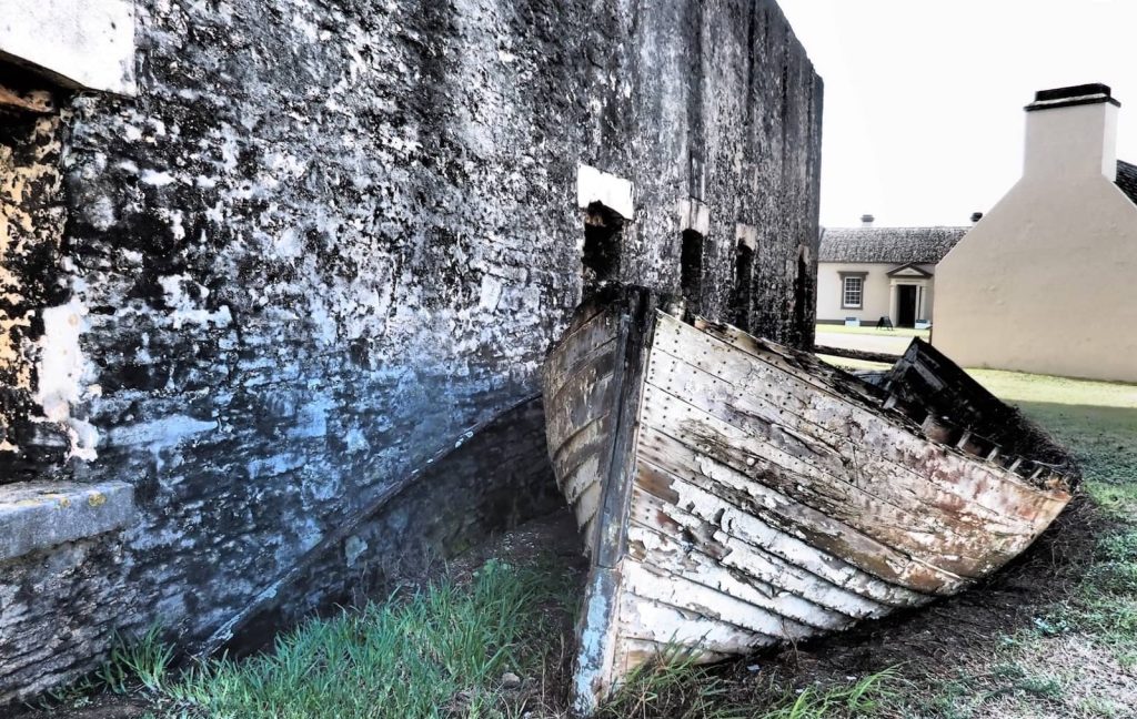 A weathered wooden boat rests against the mossy stone wall of the historic Crankmill on Norfolk Island, with colonial buildings visible in the background.