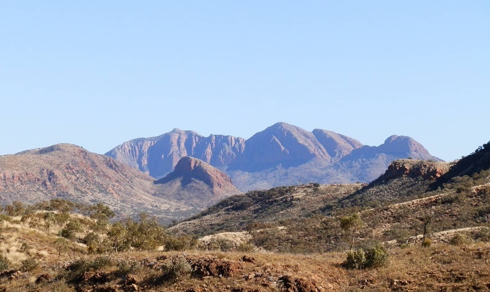 A panoramic view of the rugged West MacDonnell Ranges with Mt Sonder standing prominently in the background under a clear blue sky. The foreground features rolling, arid terrain dotted with dry shrubs and rocky outcrops.