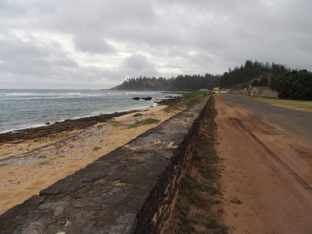 A long, weathered stone sea wall runs parallel to a coastal road on Norfolk Island, with a rugged shoreline and soft waves on the left and a forested hill and colonial buildings in the background under a cloudy sky.