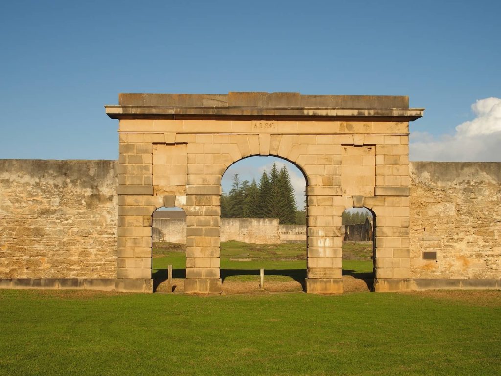 Historic sandstone prison gate dated A.D. 1847 on Norfolk Island, with three arched openings and weathered stone walls surrounding a grassy courtyard and tall pine trees in the background.