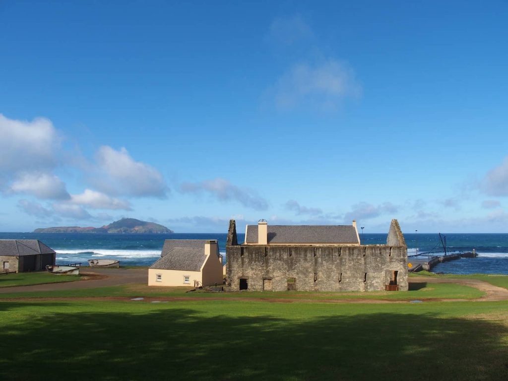 Historic stone and beige buildings sit along the coast of Slaughter Bay on Norfolk Island, with a deep blue ocean, small dock, and a distant island visible under a sunny sky.