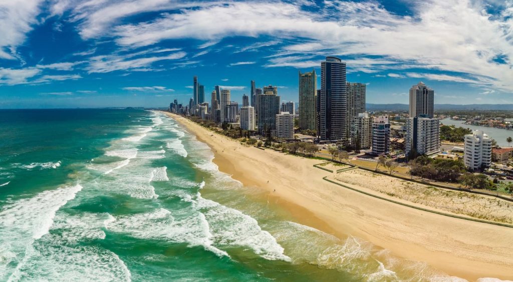 Aerial view of Surfers Paradise Beach on the Gold Coast, showcasing golden sands, turquoise waves, and high-rise buildings lining the shore. The ocean stretches into the horizon under a partly cloudy blue sky.