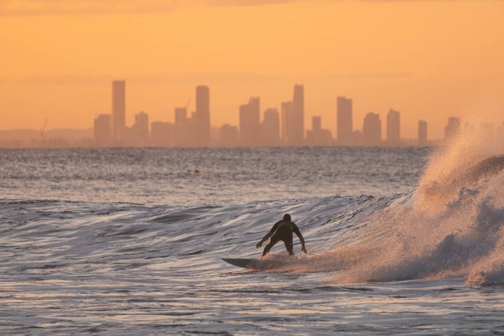 A surfer rides a wave at sunset, with the Gold Coast skyline silhouetted in the background. The golden hues of the setting sun reflect off the water, casting a warm glow over the scene. A spray of ocean mist rises as the wave crashes, framing the surfer in motion.