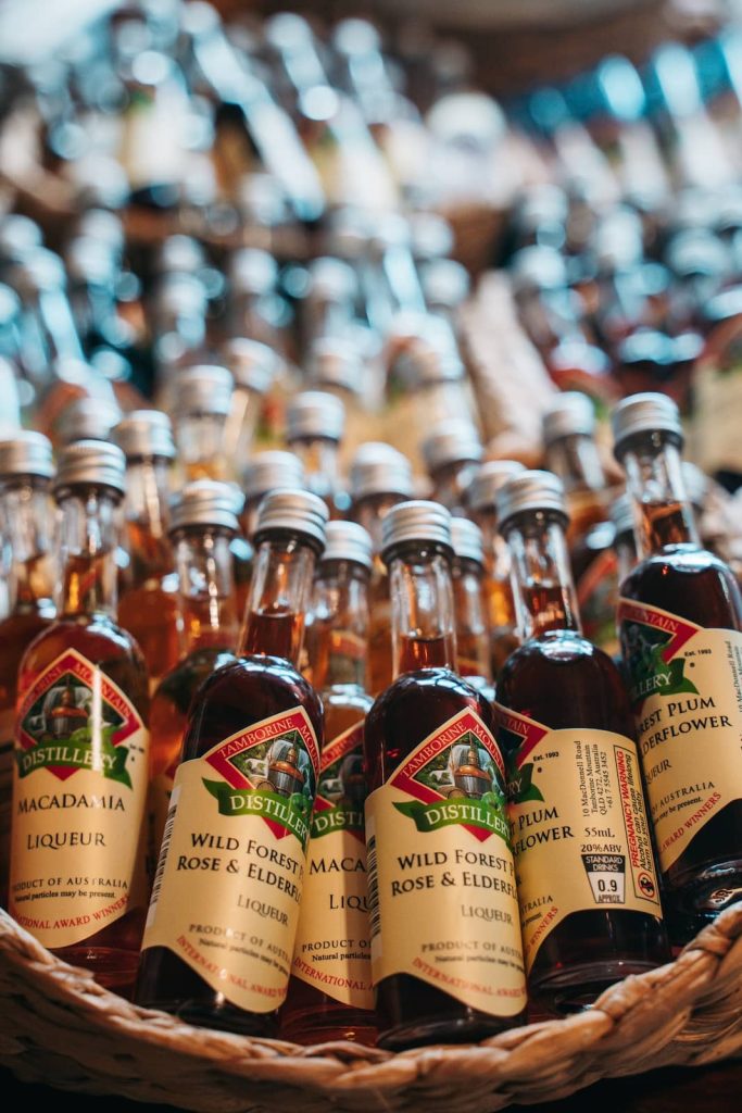 A close-up of small bottles of Tamborine Mountain Distillery liqueurs, neatly arranged in a basket. The labels showcase flavors like macadamia, wild forest plum, rose, and elderflower. The bottles have silver caps and rich amber-colored liquid inside.