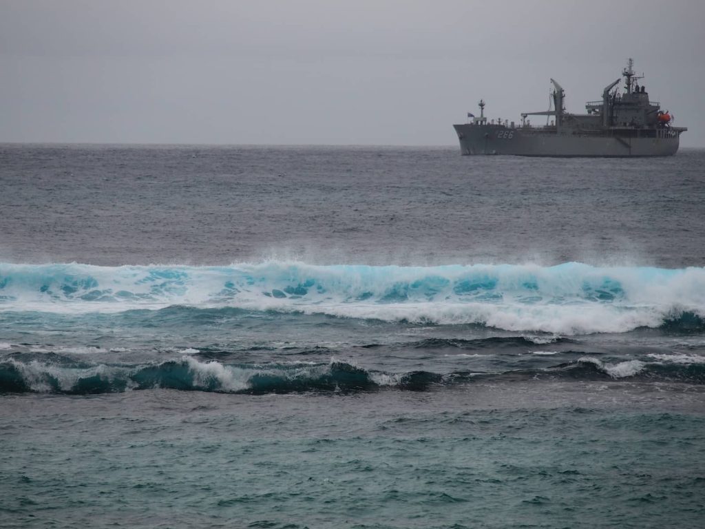 A grey naval ship, marked with the number 266, is moored off the coast of Norfolk Island, with vibrant turquoise waves breaking in the foreground under an overcast sky.