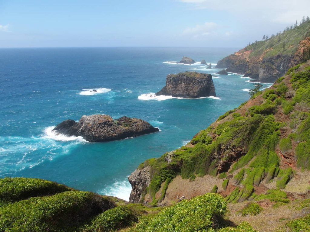 View from Captain Cook Lookout on Norfolk Island showing dramatic sea stacks rising from the turquoise ocean, with waves crashing at their bases and lush green coastal cliffs in the foreground under a bright sky.