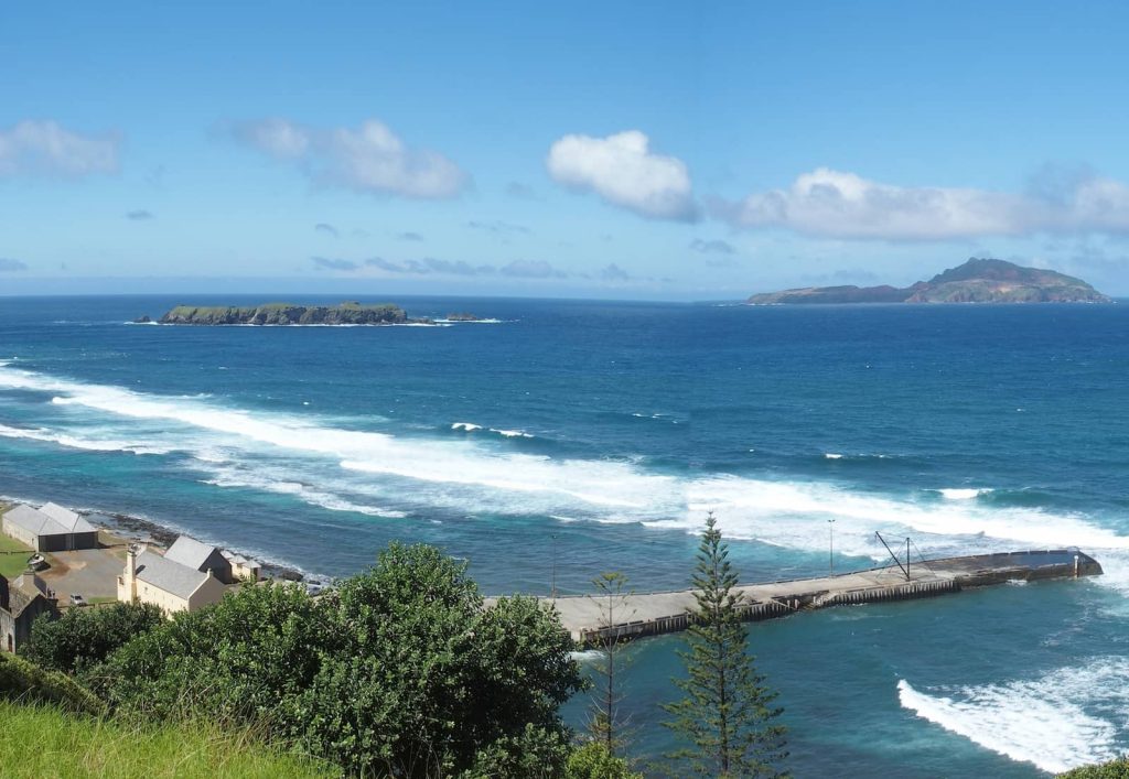 Coastal view from Flagstaff Hill Lookout on Norfolk Island, showing turquoise waves breaking along the shore, a distant rocky islet on the horizon, and heritage buildings with pale yellow walls nestled near the pier below.