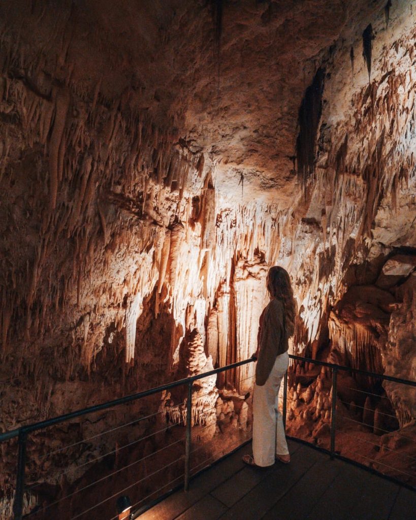 Woman standing inside a limestone cave in Margaret River, admiring dramatic stalactites and flowstone formations on a Perth to Margaret River road trip. The cavern is softly lit, highlighting natural textures.