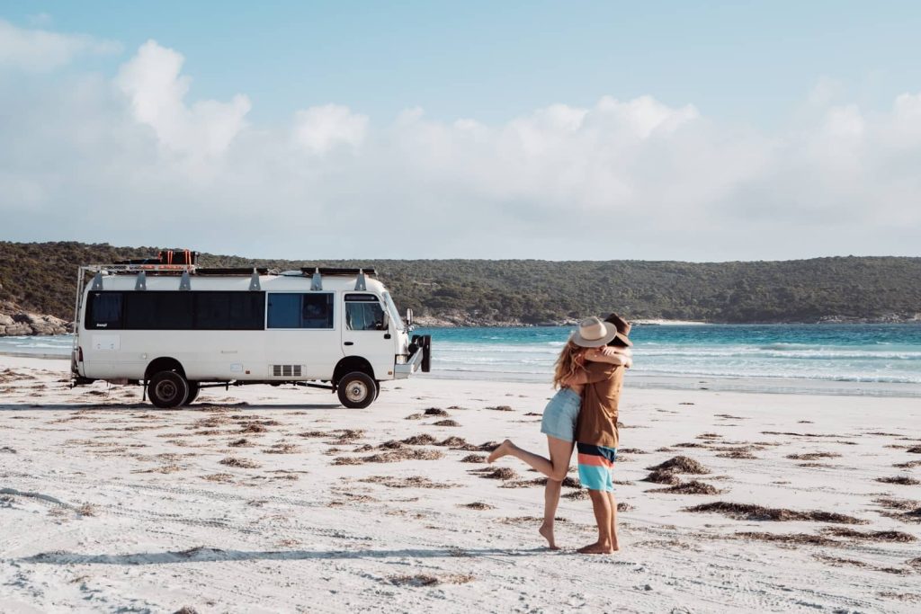 A couple embraces on the white sand at Bremer Bay with their camper parked nearby, soaking in the coastal charm of a Perth to Bremer Bay road trip.