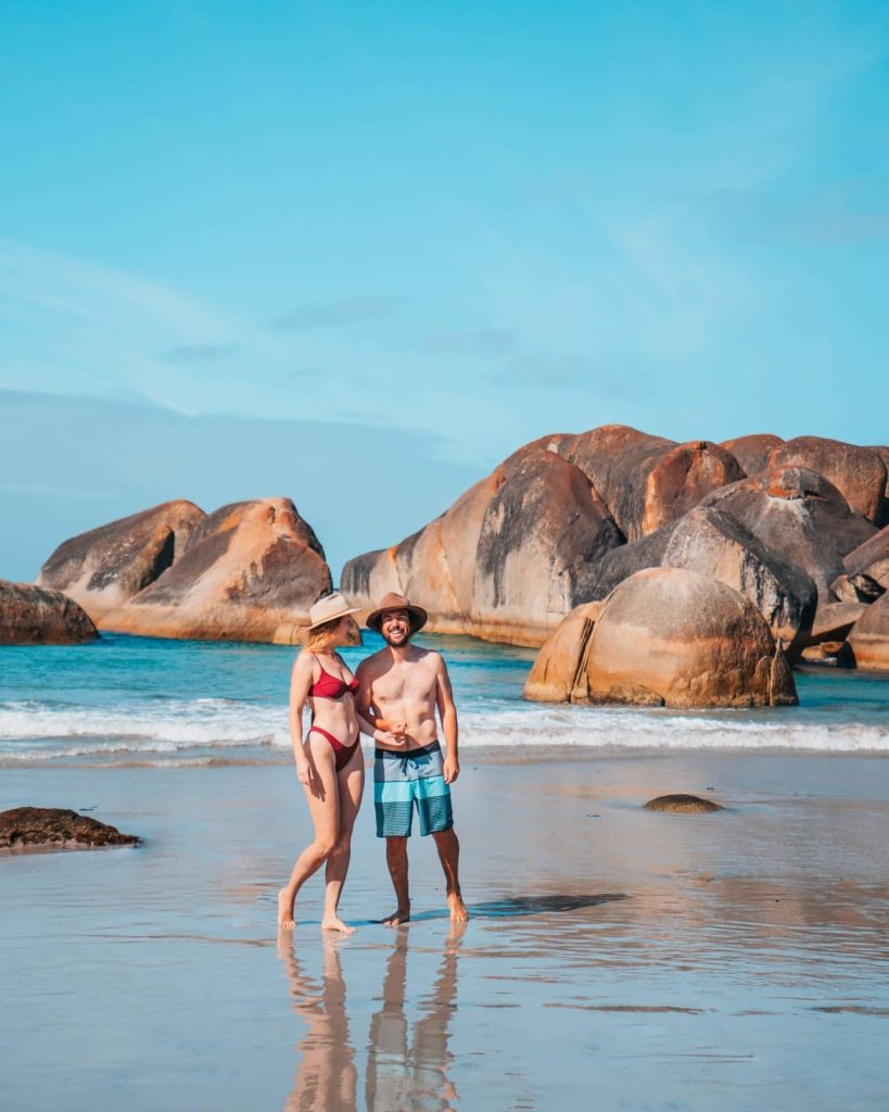 Couple standing on the wet sand in front of giant rounded boulders at Elephant Rocks in Denmark, a photogenic spot on any Perth to Albany road trip.