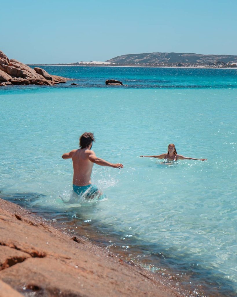 A man runs into the vibrant blue waters of Wharton Beach to join a woman swimming, showcasing Esperance’s coastal beauty not far from the Perth to Bremer Bay road trip region