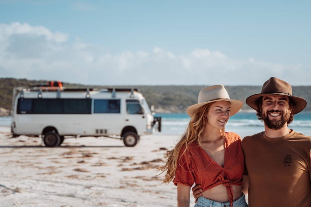 A smiling couple stands on a white sand beach with a campervan in the background, enjoying the relaxed coastal lifestyle of a Bremer Bay road trip.