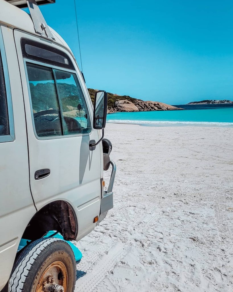 Close-up of a van parked on white sand at Wharton Beach, with vivid blue ocean and granite headlands, a relaxed stop on the Perth to Albany road trip.