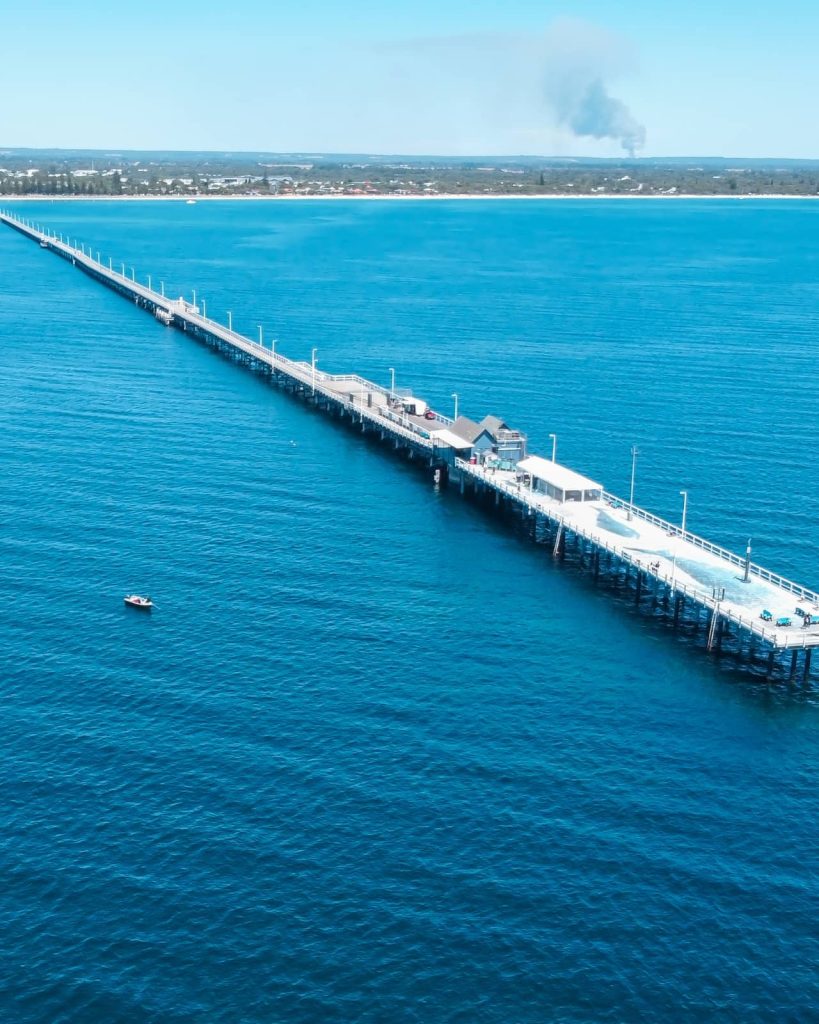 Drone shot of Busselton Jetty stretching far into the ocean with the coastline visible in the background, featured along the Perth to Margaret River road trip route. A small boat floats beside the jetty.