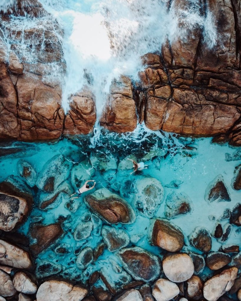 Two people relax in turquoise rock pools at Injidup Natural Spa in Yallingup, where ocean waves crash over the boulders — a scenic stop on the Perth to Margaret River road trip.