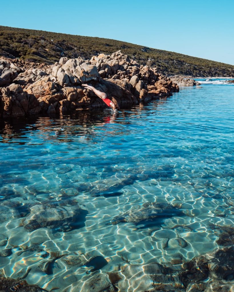 A person in red swimwear dives into the clear blue water at The Aquarium in Yallingup, surrounded by rocky shoreline, on a Perth to Margaret River road trip. The water is glassy and pristine.