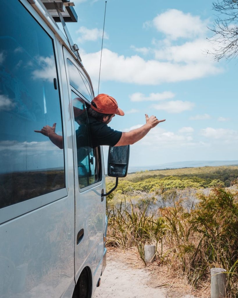 Person sticking their arm out the window of a white campervan, flashing a shaka sign with a scenic forest view in the background, symbolising freedom during a Perth to Margaret River road trip.