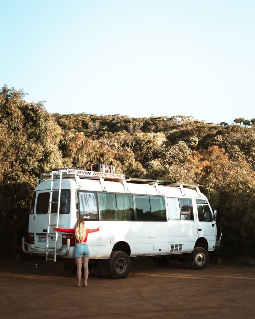 Woman in red and blue stretches her arms wide in front of a parked white campervan surrounded by trees, embracing van life on a Perth to Margaret River road trip through Western Australia.