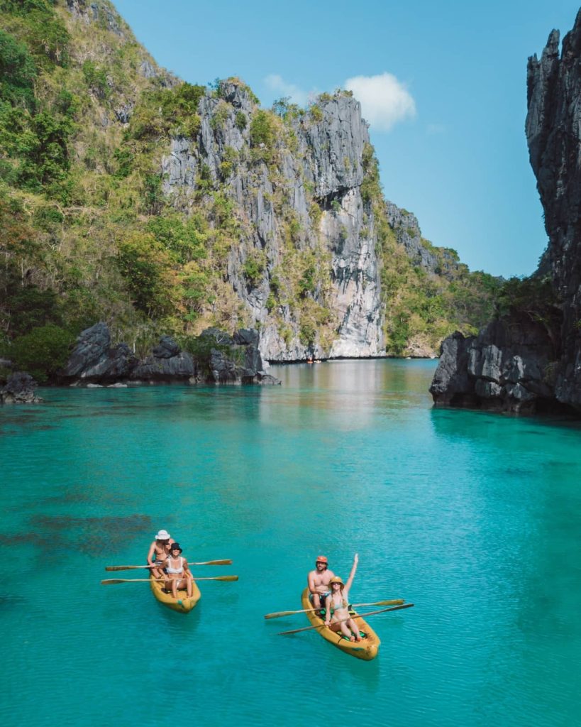 Four people paddle yellow kayaks through the turquoise waters of Big Lagoon in El Nido, Palawan. Towering limestone cliffs surround the lagoon under a bright blue sky.