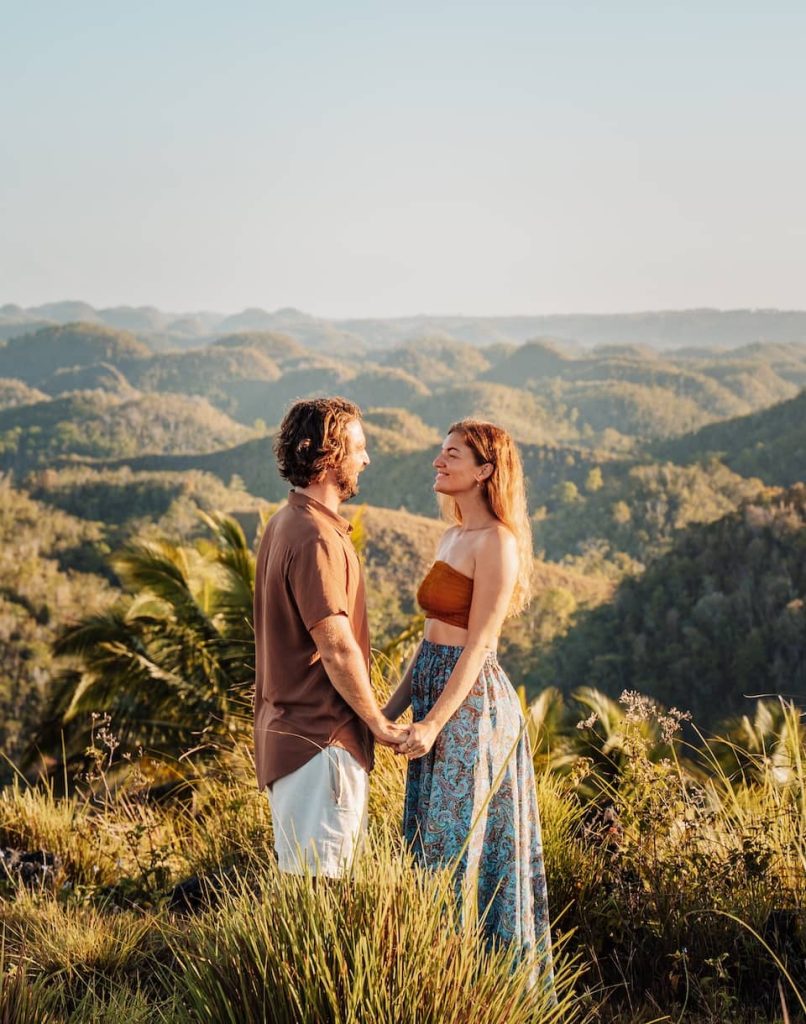 A couple holding hands and smiling at each other in a grassy hilltop overlooking the iconic Chocolate Hills in Bohol, Philippines.