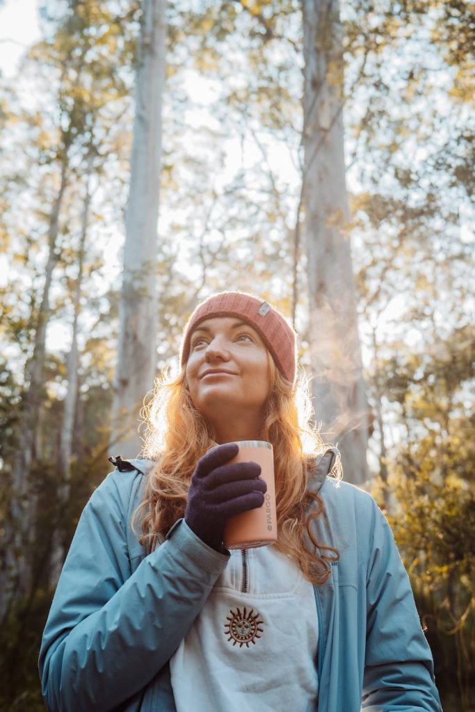 Woman wearing a beanie and gloves, holding a warm drink and standing in a forested area while camping in the Blue Mountains, NSW.