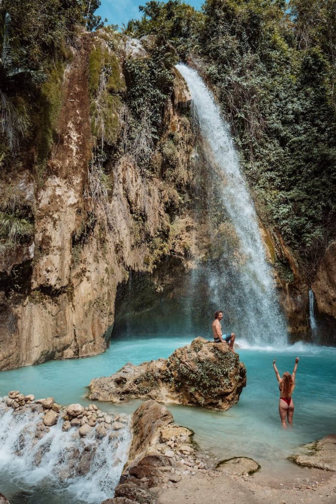 A man sitting on a rock and a woman standing in turquoise water at the base of a tall jungle waterfall in Cebu, Philippines.